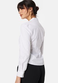 Blusa bianca a maniche lunghe realizzata in tessuto morbido, con colletto classico e design aderente, con leggeri gather sulle spalle.