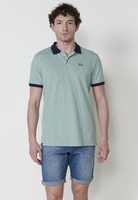 Polo košile ze světle zelené texturované tkaniny s námořnickými akcenty na límečku a rukávech. Model má na sobě srolované džínové šortky.
