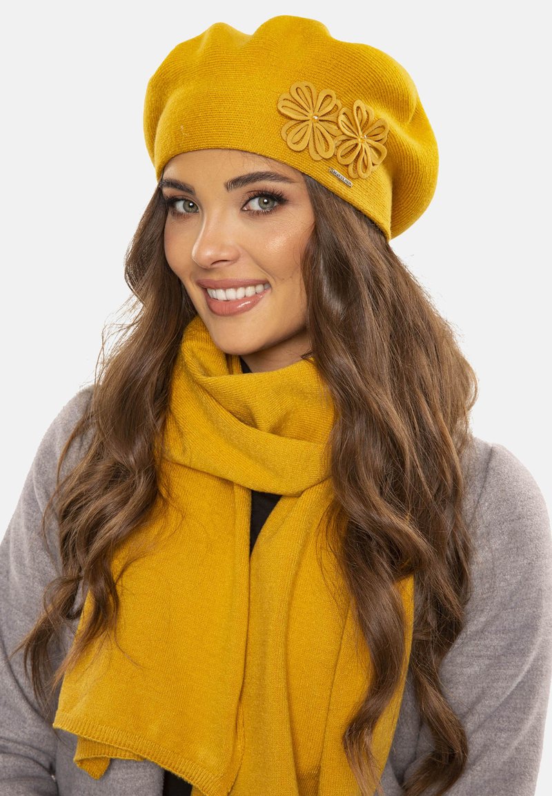Béret jaune moutarde avec un design et des détails en appliqué floral. Accompagné d'une écharpe assortie jaune moutarde, le tout en tissu doux.