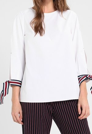 Witte top met driekwart mouwen en gestreepte boorden. De boorden hebben rode, marineblauwe en witte strepen. Gecombineerd met marineblauwe gestreepte broek.