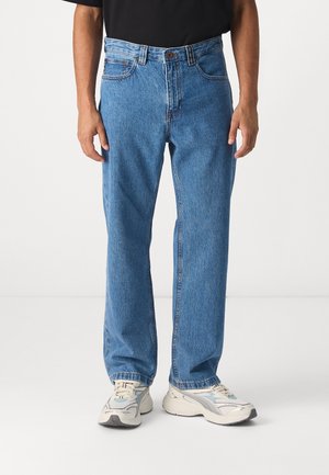 Man die een rechte-fit blauwe spijkerbroek met klassiek ontwerp met vijf zakken draagt en witte sneakers tegen een effen achtergrond.