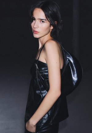 Mlajša ženska s kratkimi temnimi lasmi, oblečena v črno usnjeno opanko brez naramnic in s torbo čez ramo, pozira na temnem ozadju.