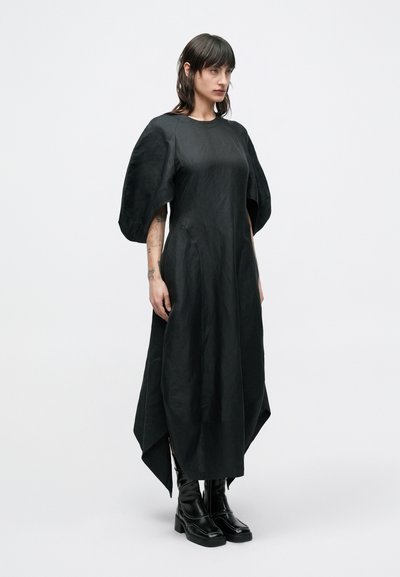 Μαύρο φόρεμα με φουσκωτά μανίκια, ασύμμετρο τελείωμα και λείο, υφασμάτινο ύφασμα με υφή. Φορεμένο με μαύρα μποτάκια με χοντρό τακούνι.