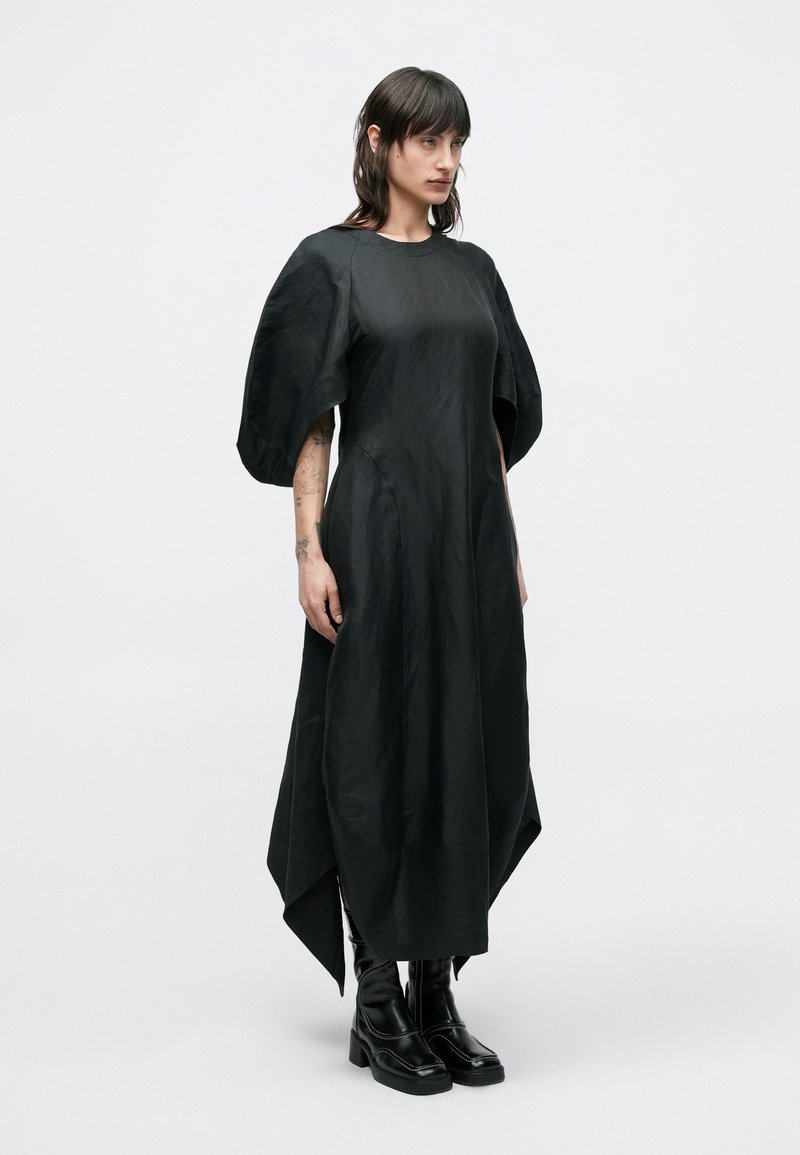 Abito nero con maniche a sbuffo, orlo asimmetrico e tessuto liscio e strutturato. Indossato con stivali neri con tacco spesso.