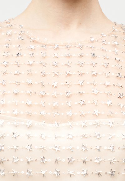 Beżowa przezroczysta tkanina zdobiona srebrnymi, metalicznymi cekinami w kształcie gwiazdek oraz małymi okrągłymi ozdobami, z okrągłym dekoltem i fakturowaną powierzchnią.