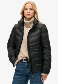 Padded black jacket with high collar, zip closure, and an emblem on the left sleeve, worn over a beige knit sweater and blue jeans.