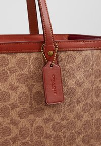 Sac fourre-tout Coach à motif marron avec des poignées en cuir et une étiquette en cuir attachée par une chaîne en laiton.