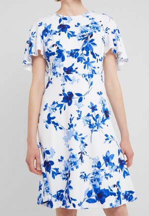 Robe blanche longueur genou avec imprimé floral bleu et manches courtes volantées portée par une personne debout.