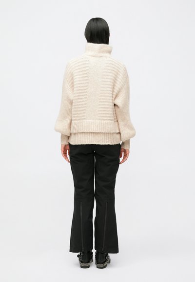 Kraftig lysebeige sweater med høj krave, ribbet tekstur og sidesømme, kombineret med sorte flarebukser med lynlåse ved manchetterne.