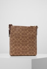 Brązowa kanwasowa torba listonoszka w kwadratowym kształcie z powtarzającym się wzorem logo, złotymi akcentami i teksturowaną powierzchnią oraz zamknięciem na zamek.