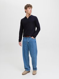 Mann trägt dunkelblauen Pullover, hellblaue Jeans, beige Schuhe und braunen Gürtel, steht mit einer Hand in der Tasche und schaut nach rechts.