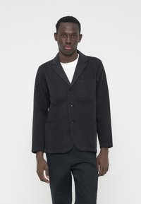 Juodas medvilninis švarkas su įpjauta apykakle, trim mygtukais ir dviem priekine kišenėmis. Dėvimas ant balto marškinio, pasižymi laisvu siluetu.