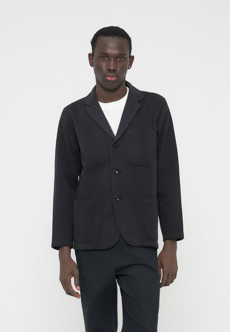 Juodas medvilninis švarkas su įpjauta apykakle, trim mygtukais ir dviem priekine kišenėmis. Dėvimas ant balto marškinio, pasižymi laisvu siluetu.