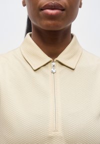 Κοντινό πλάνο του κάτω μέρους του προσώπου και του λαιμού ενός ατόμου που φοράει μια μπεζ υφασμάτινη μπλούζα με φερμουάρ και μυτερό γιακά.