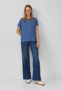 Blauwe T-shirt met korte mouwen en ribbeltextuur, gecombineerd met wijde donkerdenim jeans en witte schoenen met een crème zool. Neutrale achtergrond.