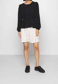 Blusa peplum nera con maniche lunghe, vita arricciata e scollatura rotonda abbinata a pantaloni corti crema e scarpe slip-on nere e chunky.