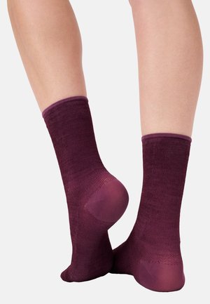 Ein Paar Beine, die mittellange, dunkelviolette, strukturierte Socken mit verstärkten Fersen und Zehen tragen, vor einem schlichten Hintergrund gezeigt.