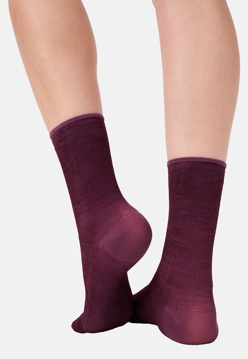 Un paio di gambe che indossano calze testurizzate viola scuro, a metà polpaccio, con talloni e punte rinforzati, mostrate su uno sfondo neutro.