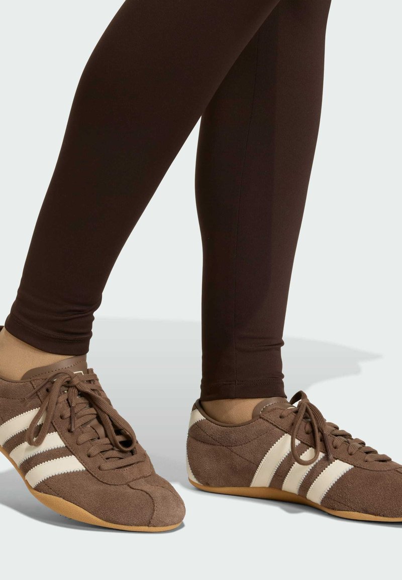 Sneaker in suede marrone con strisce bianche, lacci piatti e suola in gomma, abbinati a leggings marroni aderenti.