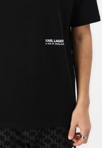 Črna bombažna majica s kratkimi rokavi, na levi strani s belim napisom "KARL LAGER" in "21 RUE ST-GUILLAUME". Preprost dizajn.