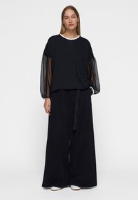 Zwarte lange mouw top met doorzichtige mesh pofmouwen, gecombineerd met zwarte wijde broek. De outfit heeft een witte geribbelde halslijn en een stoffen riem.
