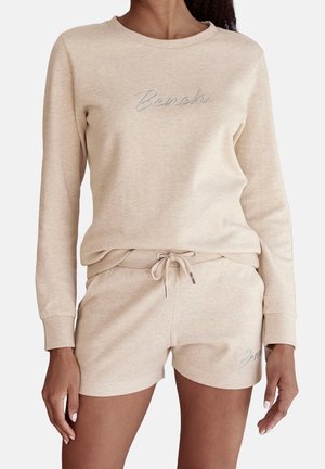 Kvinde iført beige langærmet sweatshirt og matchende shorts med snoretræk, begge broderet med ordet "Bench" i lyse tråde.