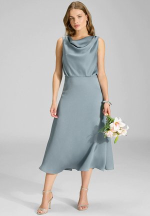 Sukienka bez rękawów z golfem w miękkim niebieskim materiale, z lejącą się spódnicą o długości midi. Modelka trzyma bukiet kwiatów. Srebrne sandały na obcasie z odkrytymi palcami.