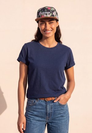 Donna sorridente che indossa un berretto a stampa floreale, una maglietta blu scuro con maniche arrotolate, una cintura marrone e jeans blu, in piedi davanti a uno sfondo beige.