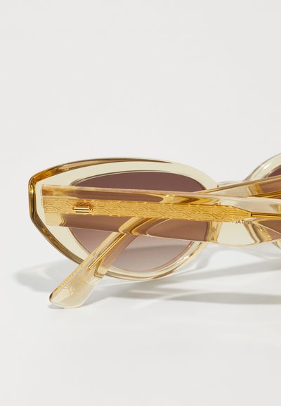 Saulesbrilles ar caurspīdīgi zeltītām rāmī, brūniem gradienta objektīviem un teksturētām dzeltenām akcentiem uz tempļiem. Eleganta, ovāla forma.