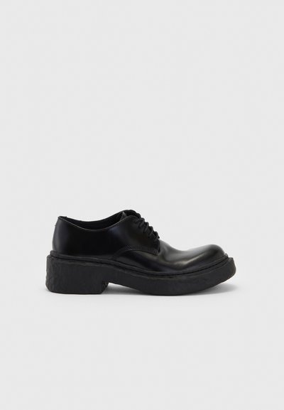 Black leather lace-up shoe with a rounded toe and a thick, textured black outsole for added height and stability. Smooth finish overall.