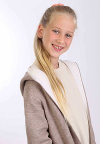Braune Kapuzenjacke mit weißem Innenfutter, weiche Textur, kombiniert mit einem hellbeigen Oberteil. Lange blonde Haare, frisiert mit einem Pfirsich-Haarband.