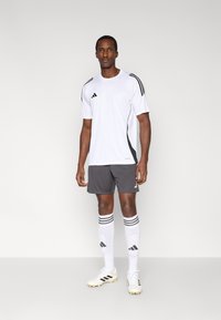 Weißes Sport-T-Shirt mit schwarzen Akzenten und drei Streifen, kombiniert mit grauen Shorts, weißen Socken mit Streifen und weißen Fußballschuhen.