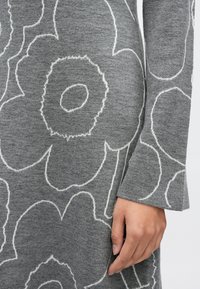 Ruka opírající se o šedou látku s bílým květinovým vzorem v obrysech, zobrazující část dlouhého rukávu a šaty nebo kabát.