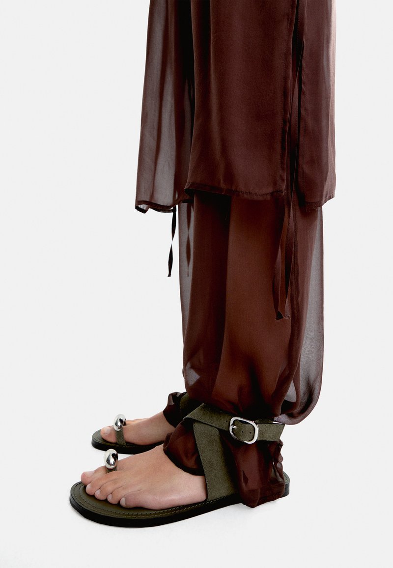 Brązowe, przezroczyste materiałowe spodnie na oliwkowych sandałach z klamrą i srebrnymi akcentami. Sandały mają płaską podeszwę i otwarty czubek.