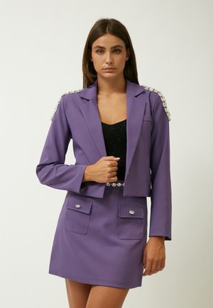 Blazer viola con spalline decorate, design aderente, due tasche frontali. Abbinato a un top nero e a una gonna strutturata coordinata.