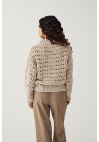 Pikkade lokkis juustega naine, kes kannab beežikat avatud kudumiga kampsunit ja helepruune pükse, seisab seljaga vaatajale ainult lihtsa taustaga.