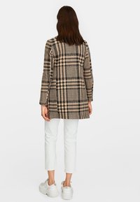 Ruudullinen pitkä takki beige ja musta, valmistettu kudotusta kankaasta, jossa on suora leikkaus, pitkät hihat ja takakappaleen halkio, yhdistetty valkoisiin housuihin.