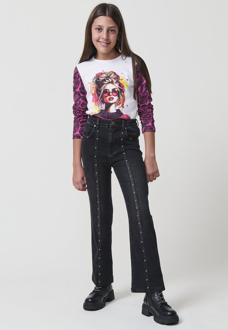 Chica de pie sonriendo, vestida con pantalones negros con tachuelas, botas negras y una camiseta blanca con mangas rosas y un gráfico de una mujer con gafas de sol.