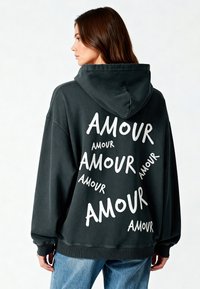 Hoodie oversize nero con testo bianco "AMOUR", dotato di cappuccio con coulisse, polsini a coste e texture di stoffa morbida.