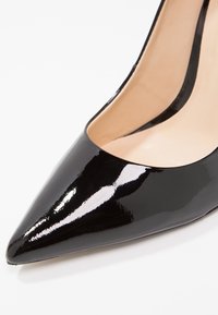 Chaussure à talon aiguille en cuir verni noir avec un bout pointu et un décolleté bas, présentant une surface lisse et brillante ainsi qu'un intérieur beige.