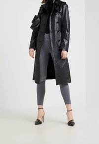 Trench coat de cuero sintético negro con botones, cuello y detalles de manga rizada. Lleva encima unos jeans ajustados grises y tacones negros.