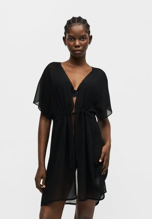 Femme aux cheveux tressés portant une robe noire transparente nouée à la taille, révélant un soutien-gorge en dentelle noire en dessous, se tenant devant un fond uni.