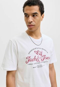 Balta trumpa rankovė t-marškiniai su raudonu spaudiniu, kuriame yra logotipas ir tekstas "Jack & Jones", su retro stiliaus akcentais ir apvaliu kaklu.