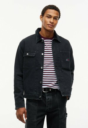 Ung mand i sort denimjakke over en striped vinrød og hvid skjorte, iført sorte bukser med den ene hånd i lommen, lys baggrund.