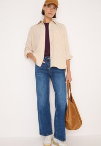 Camicia di velluto a coste beige, top viola intenso, jeans larghi blu, sneakers chiare con accenti blu e borsa a spalla in suede marrone.
