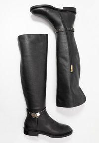 Botas negras de cuero por encima de la rodilla con un acabado texturizado, que cuentan con acentos de hardware dorado y un cierre de cremallera lateral. Diseño de suela plana.