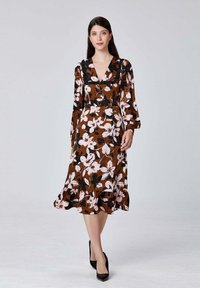 Robe à imprimé floral marron avec des manches longues, un décolleté en V et un ourlet froncé. Fleurs blanches et roses, avec une texture de tissu lisse. Escarpins noirs.