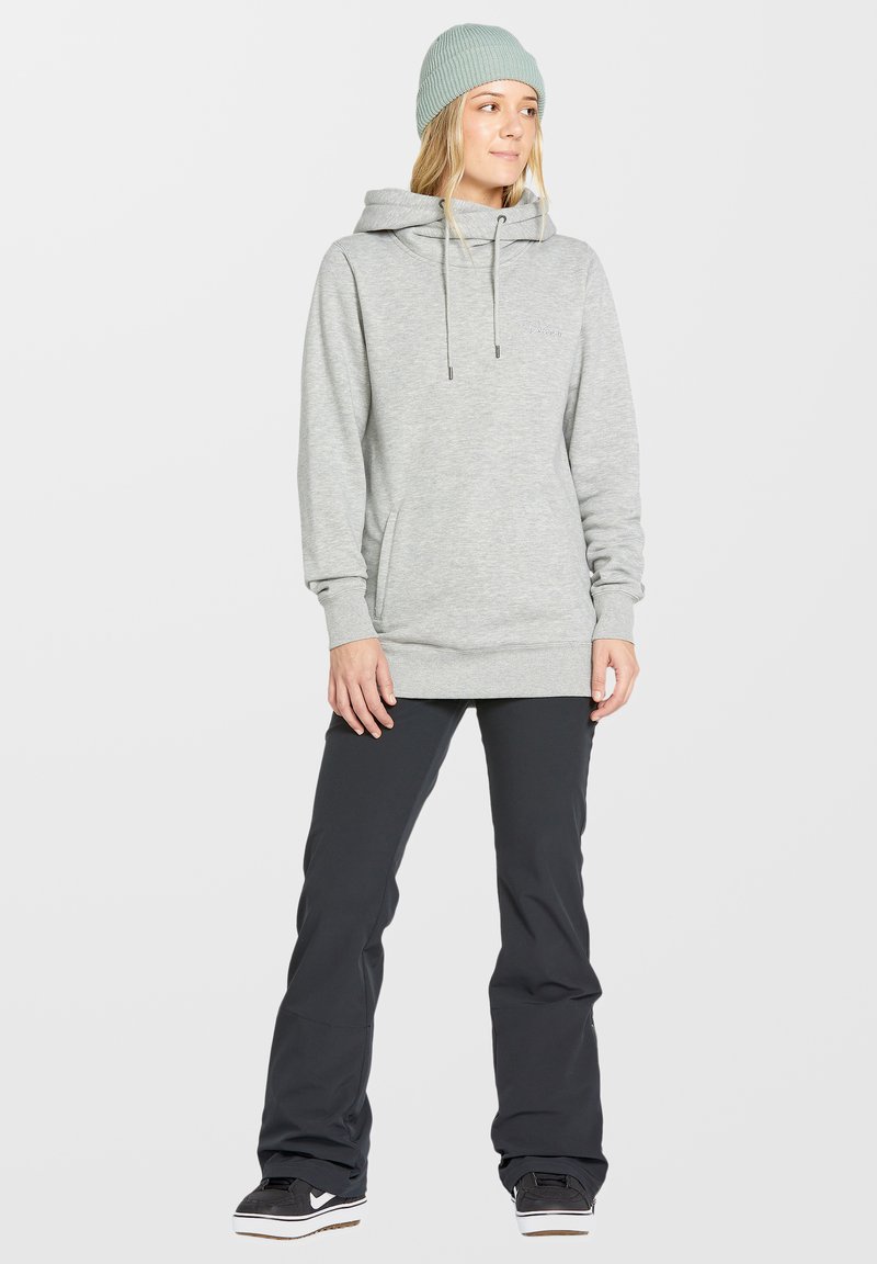 Sudadera con capucha de color gris claro, con un bolsillo frontal, cordones y un ajuste relajado, combinada con pantalones de nieve negros y zapatos negros.