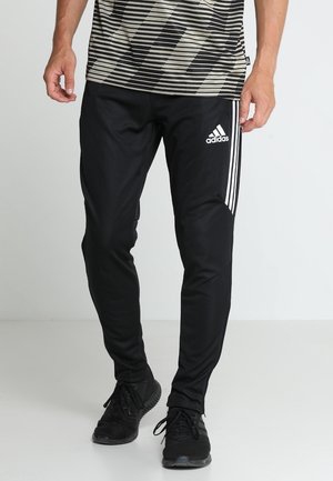 Mężczyzna ubrany w czarne spodnie dresowe Adidas z białymi paskami i logo, w połączeniu z czarnymi sneakersami i koszulą w wzory.