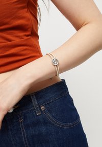 Pulsera delgada de color crema con un detalle circular de plata. La pulsera presenta una textura suave y un diseño minimalista, llevada en una muñeca con denim.